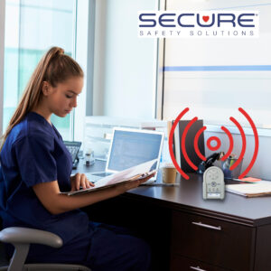 Nurse sitting at a desk reading a chart. The Secure Wireless Alarm Monitor is on the desk alerting the nurse of unsafe patient movement.